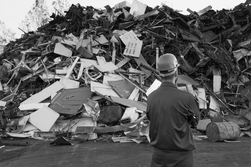 A man wearing a safety helmet and dark work clothing stands with his back to the camera, observing a large pile of mixed waste materials and discarded objects. The waste includes broken appliances, various plastics, metal debris, and cardboard, forming a dense, untidy heap on a paved surface, likely within a waste management or recycling site near Feltham. The background reveals a clear sky and some trees, suggesting an outdoor environment. The scene is typical of waste collection or rubbish removal activities, highlighting the need for professional rubbish disposal services such as those provided by Business Waste Removal Feltham. The materials vary in texture and color, with some items appearing smooth and others jagged or damaged, indicating they are ready for sorting or transportation. The overall setting emphasizes industrial waste management in the local area, with the individual in protective gear overseeing a substantial accumulation of refuse, ready for removal or processing.