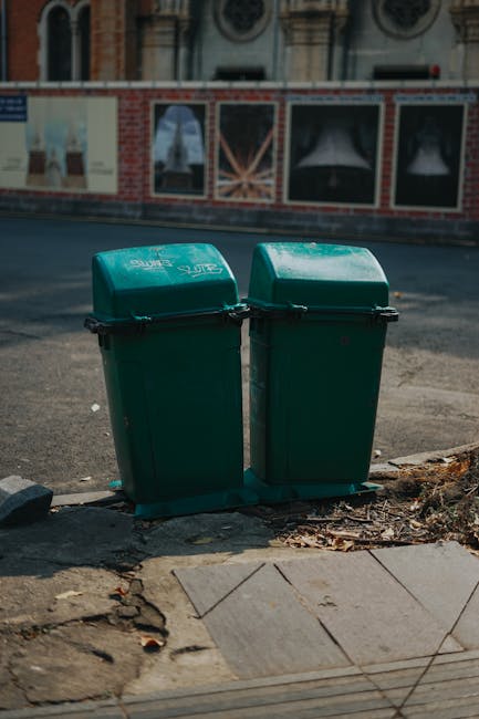 Two green plastic recycling bins with black clasps and slightly curved lids are positioned side by side on a paved urban sidewalk. The bins have a matte finish and are placed directly on the ground, w
